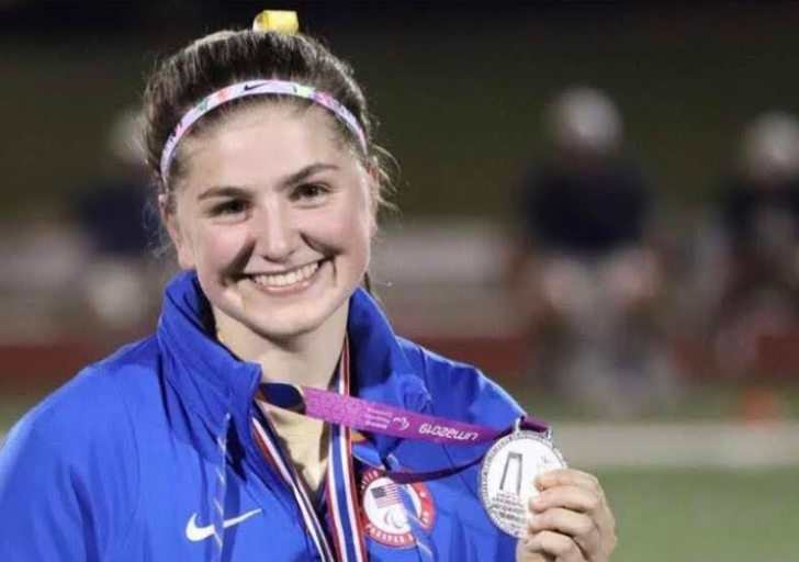 An athlete with a physical disability smiles while holding a silver medal at a track meet, wearing a blue jacket and headband under stadium lights.