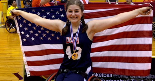 An athlete with a physical disability celebrates in a wheelchair basketball arena, wearing a USA jersey and medal while holding an American flag with both arms extended.