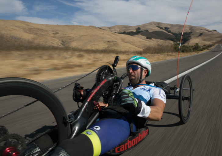 A handcyclist wearing a helmet and sunglasses rides along an open highway, powering the handcycle with a strong forward motion. Rolling hills and a blue, partly cloudy sky stretch across the background.
