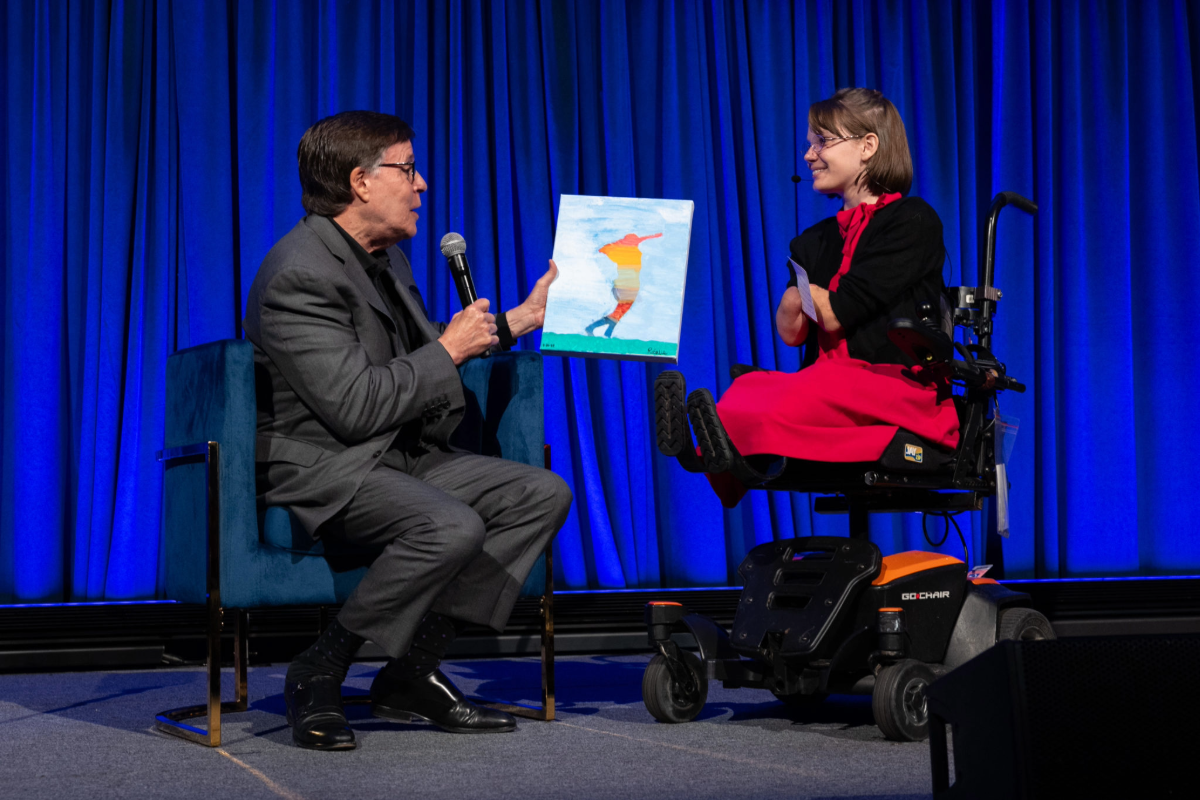 A man in a suit holds a microphone and displays a colorful painting on stage while speaking with a young woman in a power wheelchair wearing a red dress. The background is a curtain lit in blue.