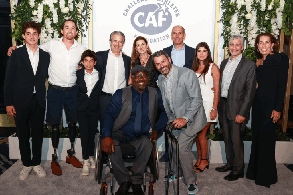 Group photo of attendees at the Challenged Athletes Foundation Heroes, Heart & Hope Gala. People in formal attire stand smiling in front of a floral backdrop with the CAF Foundation logo. Some individuals use wheelchairs and prosthetic legs.