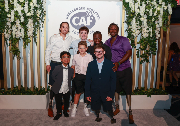 vGroup of athletes and youth dressed in semi-formal attire posing together in front of the Challenged Athletes Foundation backdrop with floral decorations. Several individuals wear prosthetic legs
