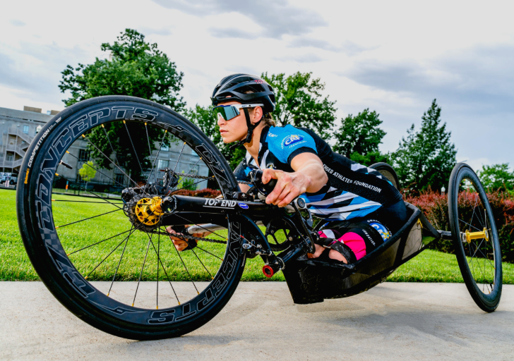 A para-cyclist races in a sleek black handcycle, leaning low to the ground while wearing a helmet and reflective sunglasses.