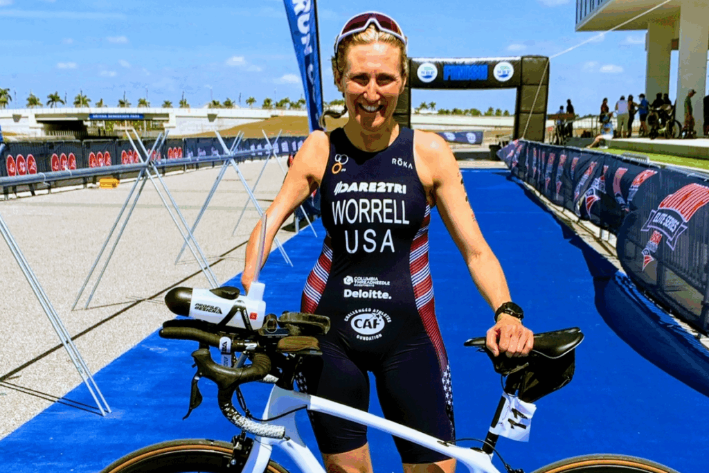 A female para-triathlete in a USA tri-suit stands smiling with her bike on a blue finish line carpet at a competition venue.