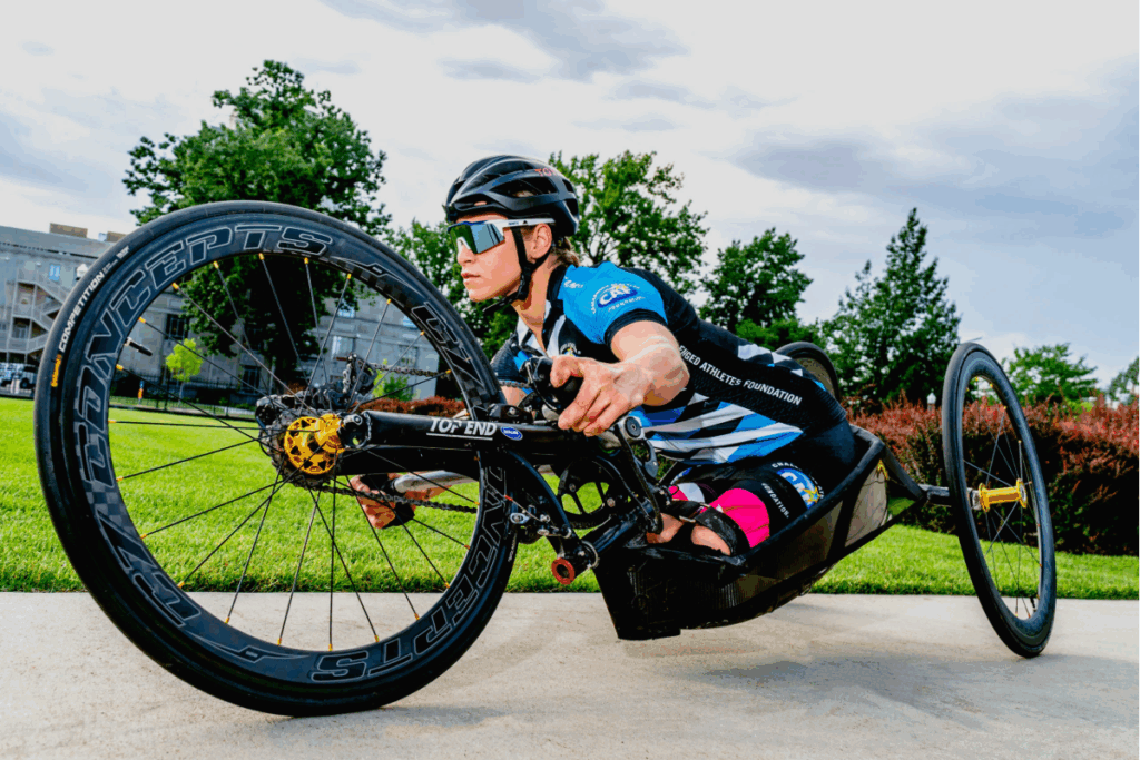 A para-cyclist races in a sleek black handcycle, leaning low to the ground while wearing a helmet and reflective sunglasses.