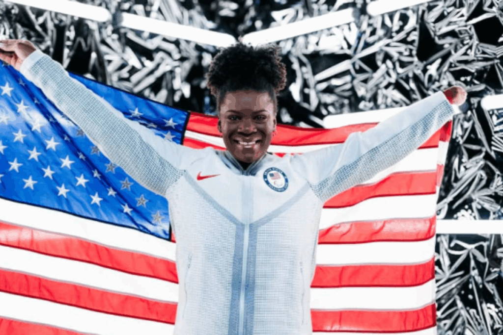 A Paralympic athlete smiles broadly while holding the American flag behind her, wearing a white Team USA jacket.