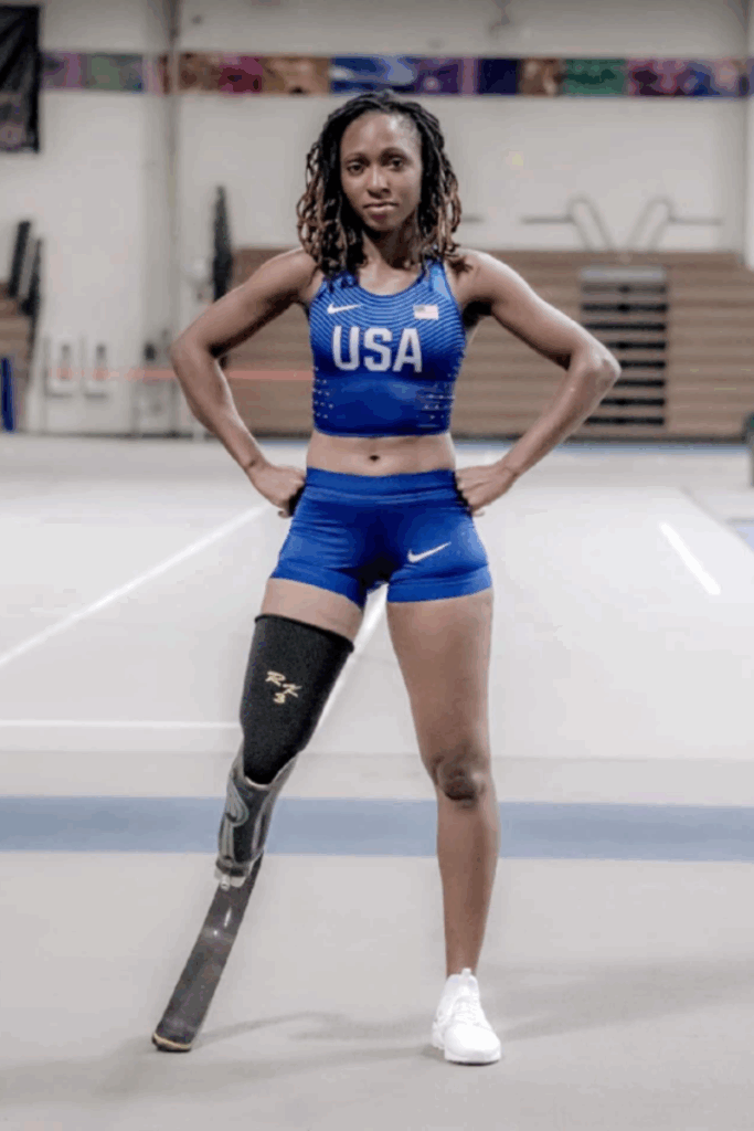 A female track and field athlete wearing a USA crop top and shorts poses confidently indoors, standing with a running blade prosthetic.