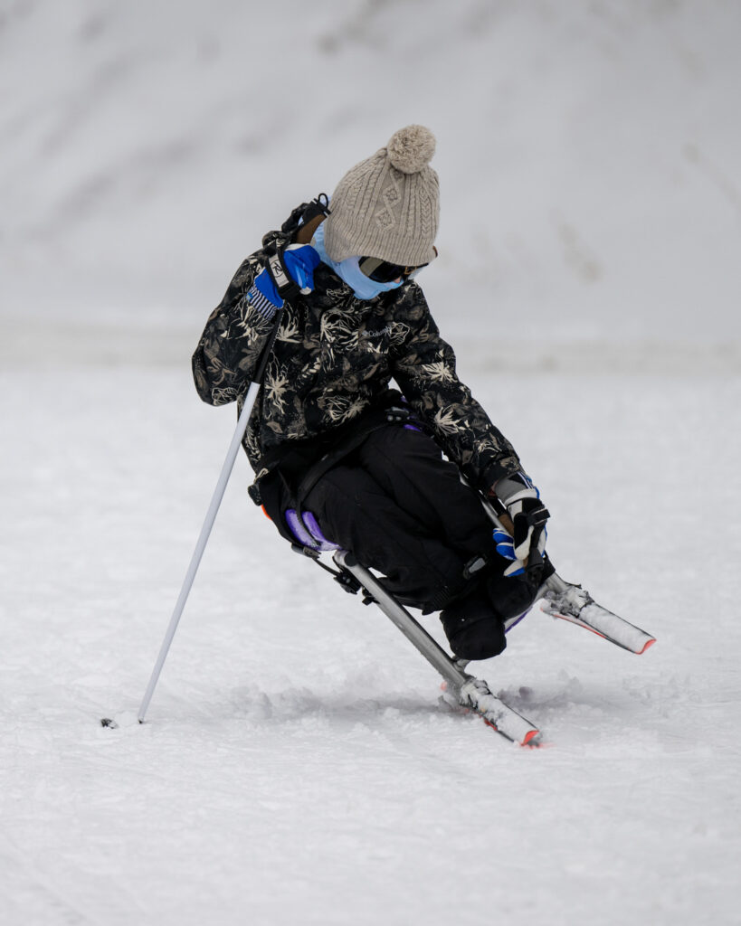 A child skiing on a sit-ski wearing winter clothing and a beige beanie.