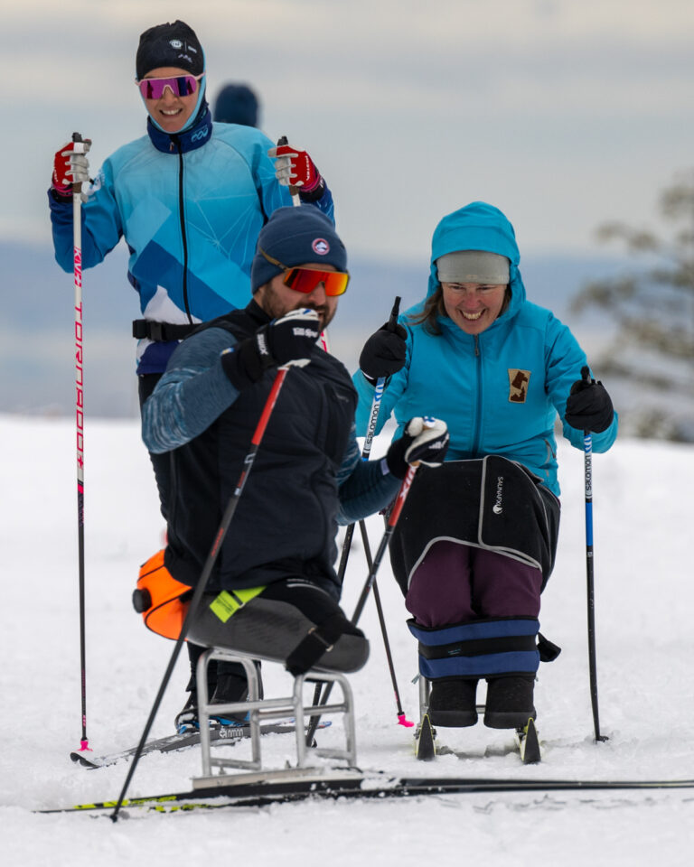 Three people skiing, two in sit-skis and one standing, on a snowy surface.