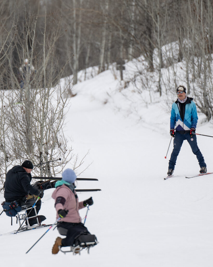 Three people skiing on a snowy path, two using adaptive ski equipment and one skiing upright with leafless trees around.