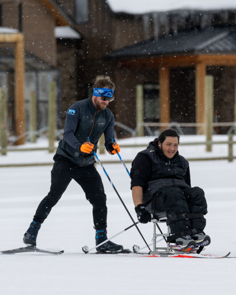 Two people in a snowy scene; one skiing, the other on a sit-ski.