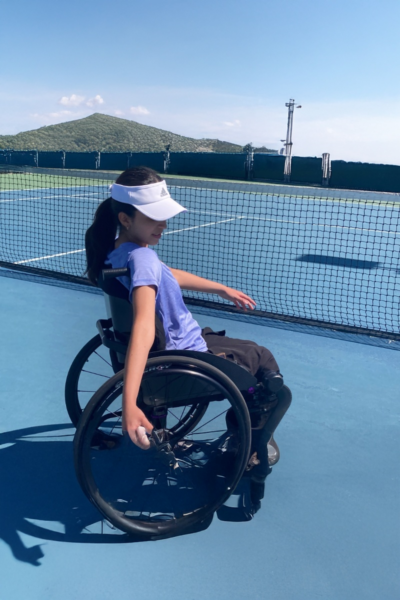A young athlete in a sport wheelchair practices on an outdoor tennis court, reaching with one arm toward the ball while wearing a visor and athletic gear under a bright blue sky.