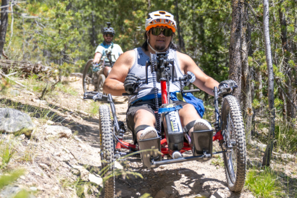 An athlete rides an off-road handcycle down a dirt trail surrounded by pine trees, wearing a helmet, sunglasses, and gloves as another rider follows behind.