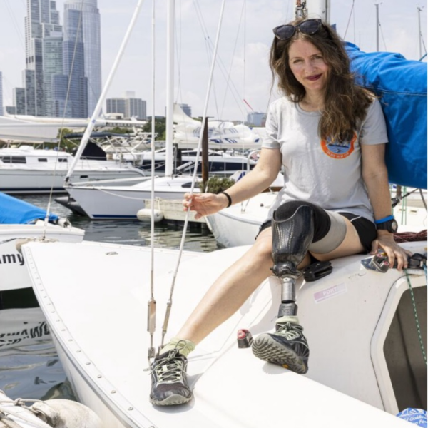 A sailor with a prosthetic leg sits on the deck of a sailboat in a marina, smiling toward the camera with city buildings in the background.
