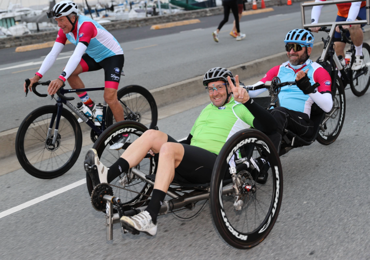 Two athletes ride handcycles alongside a cyclist on a coastal road, smiling and signaling as they move together.