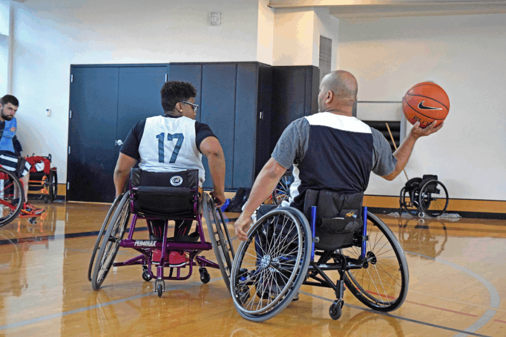 Two athletes in sport wheelchairs on a basketball court, one holding a ball and preparing to pass.