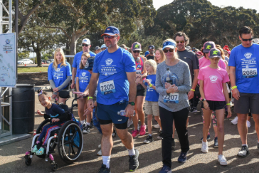 A group of participants, including wheelchair users and families, walk together under the welcome banner to kick off the 5K Walk + Roll.