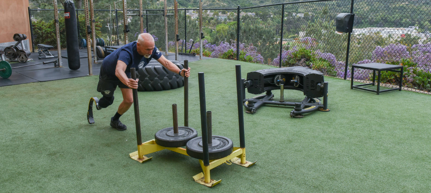 Athlete with a prosthetic leg pushes a weighted sled outdoors on turf, showcasing strength training as part of virtual participation.