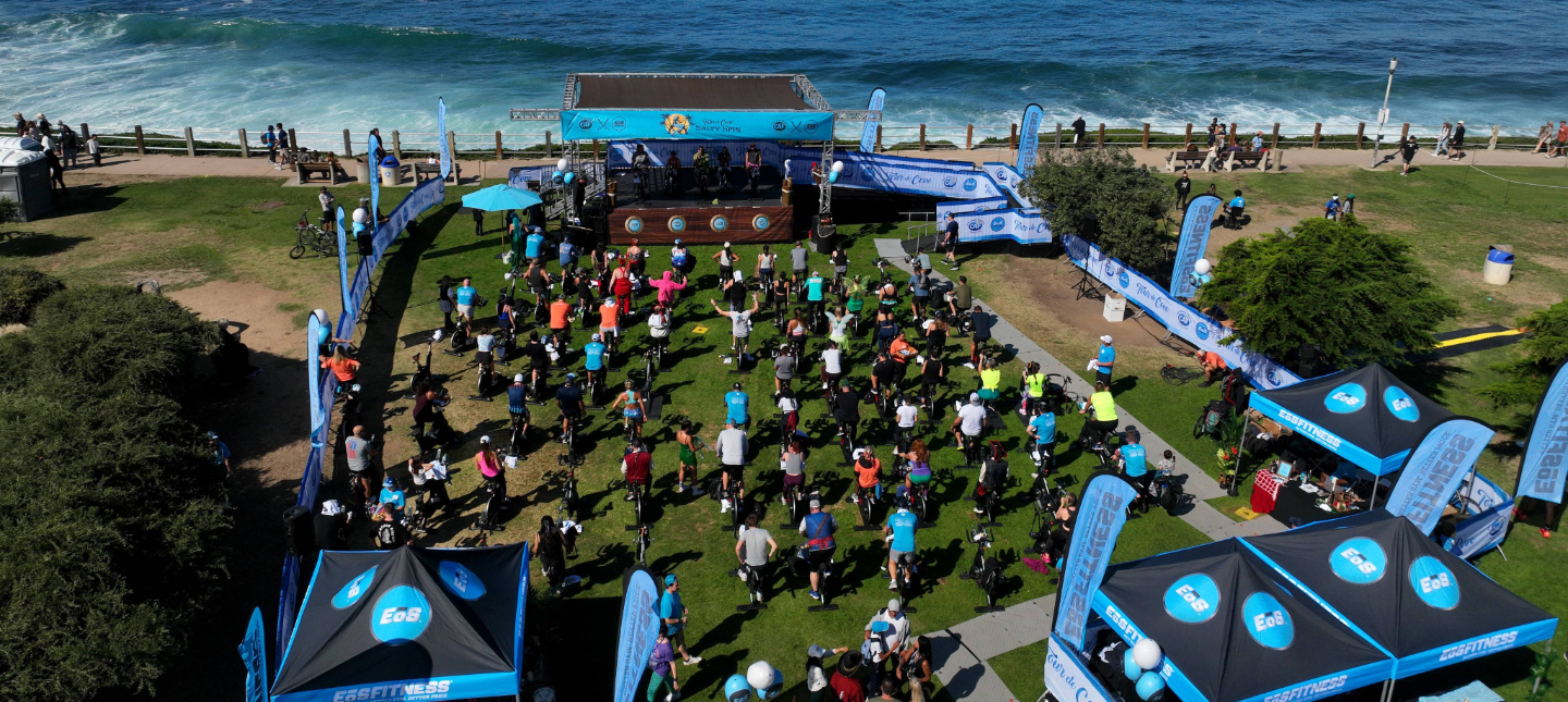 A large outdoor cycling event on a grassy bluff overlooking the ocean, with dozens of participants riding stationary bikes facing a stage and sponsor banners.