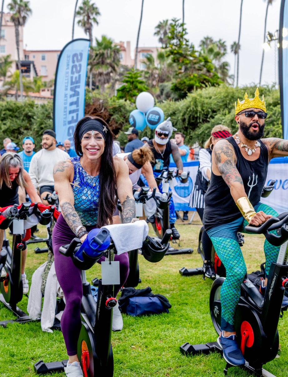 Two participants in themed costumes smile while riding stationary bikes outdoors, with other cyclists and palm trees in the background.