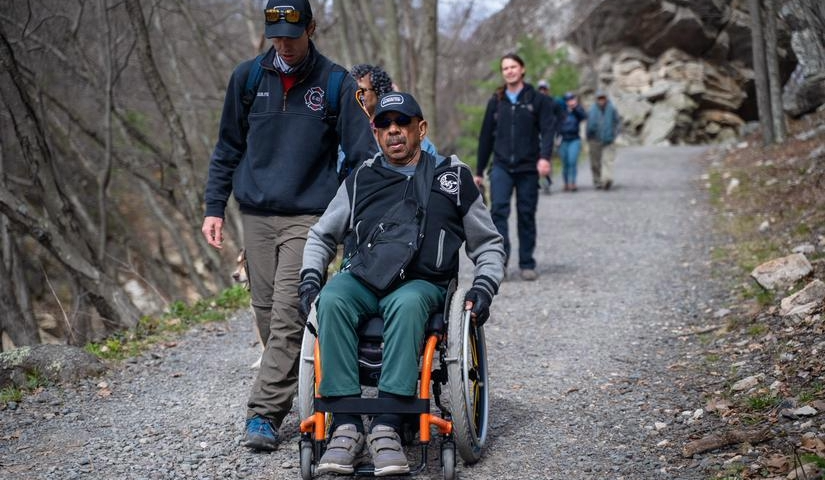 Man in wheelchair rolling next to man walking on a hiking path