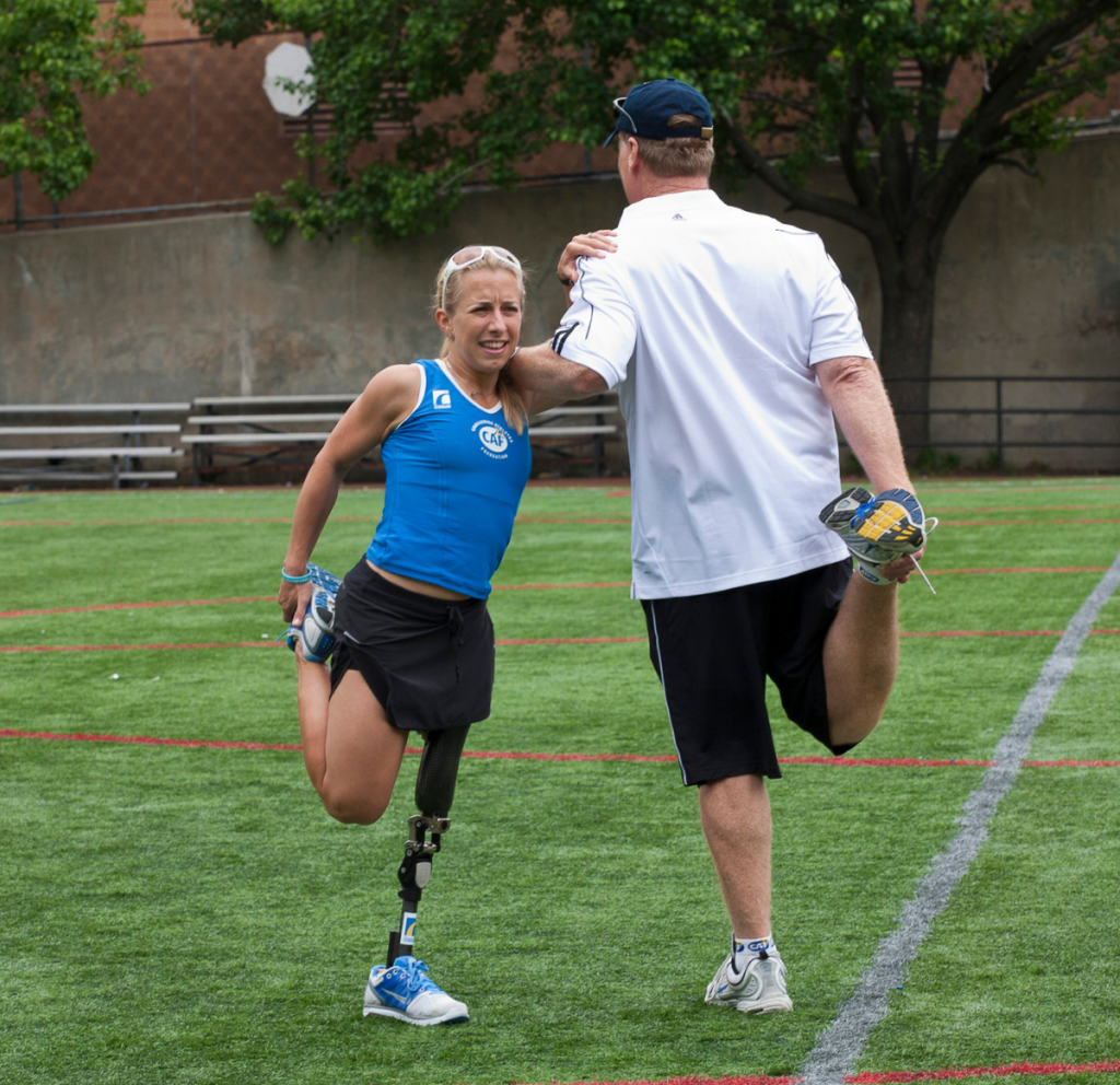 Athlete with a prosthetic leg stretches with a coach on a field.