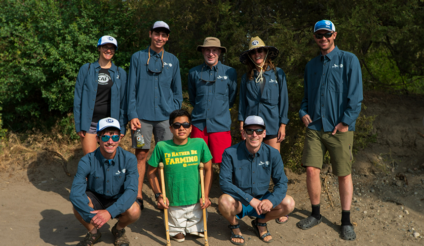 Group of volunteers in long sleeve blue shirts and one young athlete in a green shirt and crutches posing for a photo with a background of green trees.