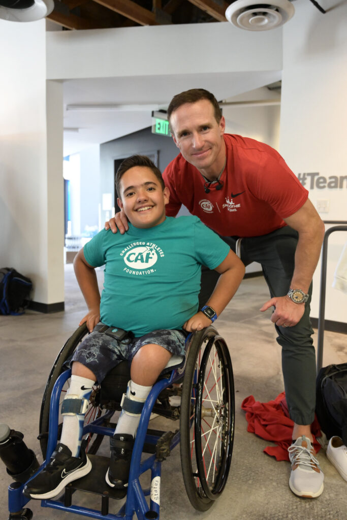 A young person in a wheelchair beside an adult, both smiling, indoors.