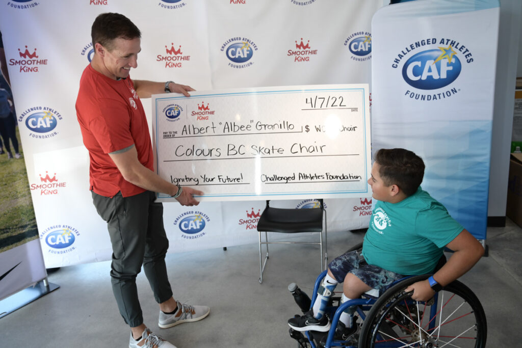 A person presents a large ceremonial check to a child in a wheelchair in front of a promotional backdrop.