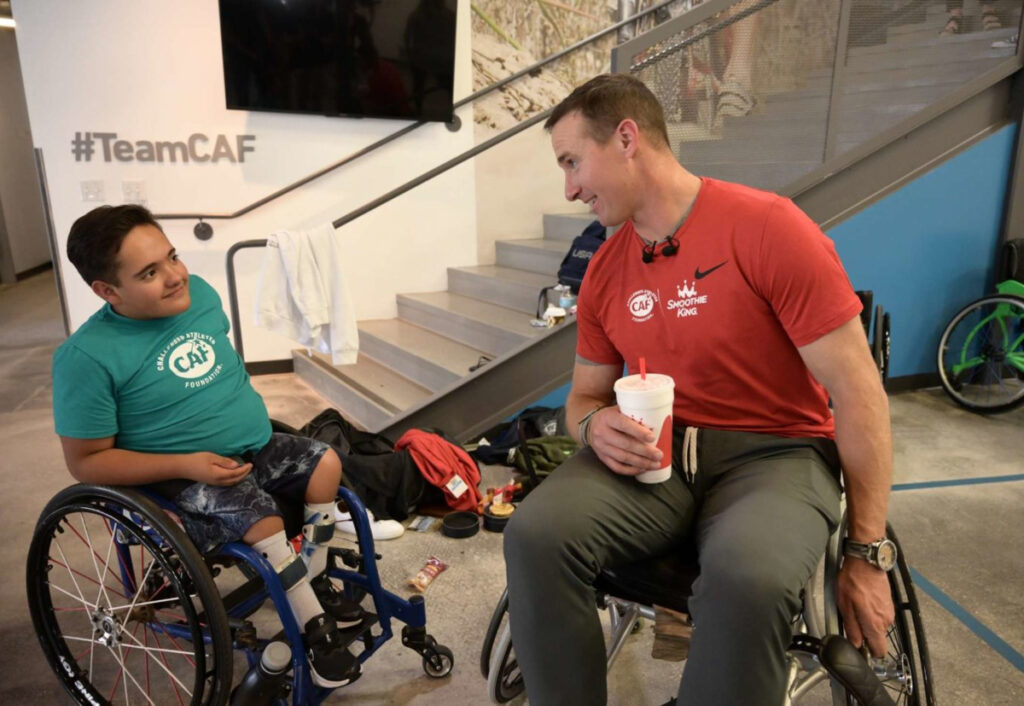 Two people in wheelchairs talking indoors, one in a teal t-shirt, the other in red.