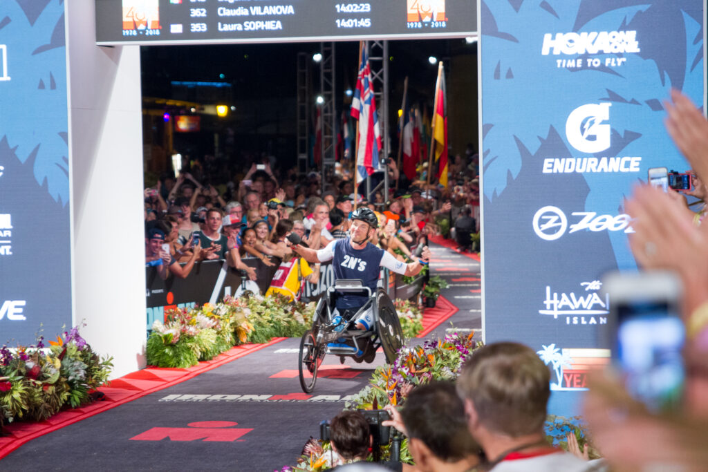 Glenn Hartrick crossing the finish line of the Ironman World Championship in Kona in a handcycle, arms raised in celebration