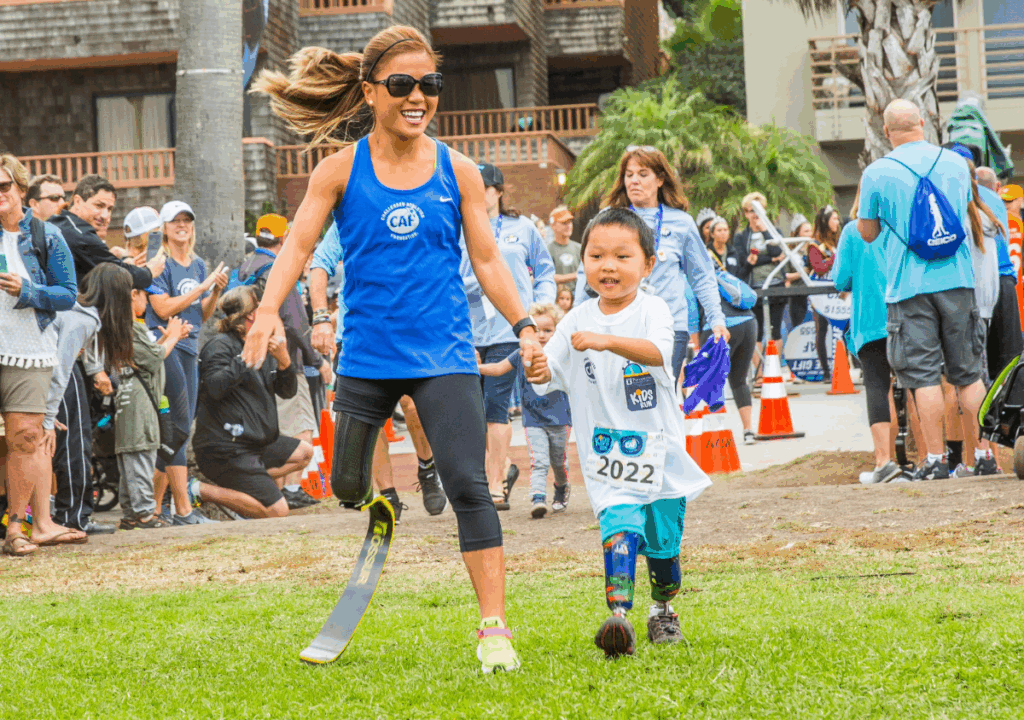 Scout Bassett holding hands and running with a young boy wearing a prosthetic leg at the San Diego Triathlon Challenge Kids Run