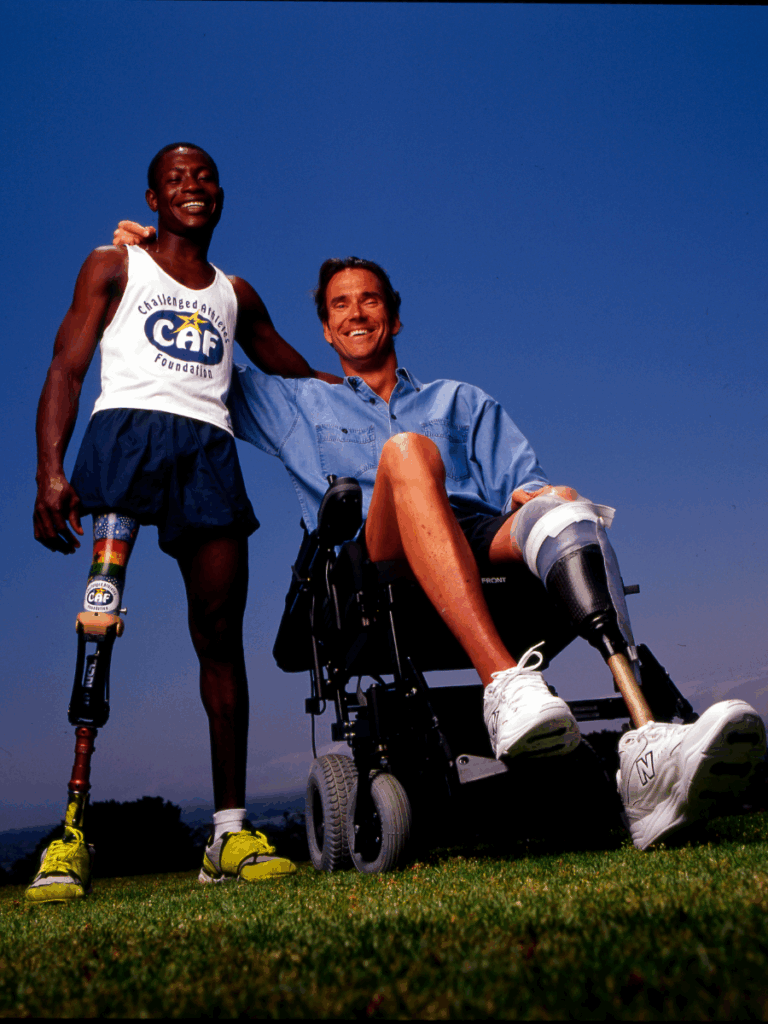 Jim MacLaren sitting in a wheelchair with another athlete, both with prosthetic legs, smiling side by side