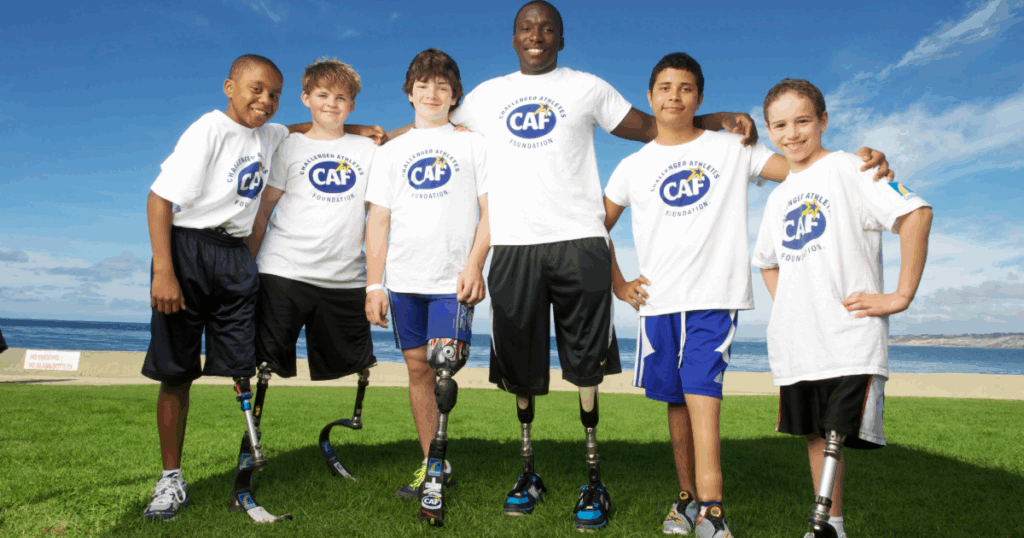 Six youth athletes, several with running blades and prosthetic legs, smiling arm-in-arm by the ocean in CAF shirts