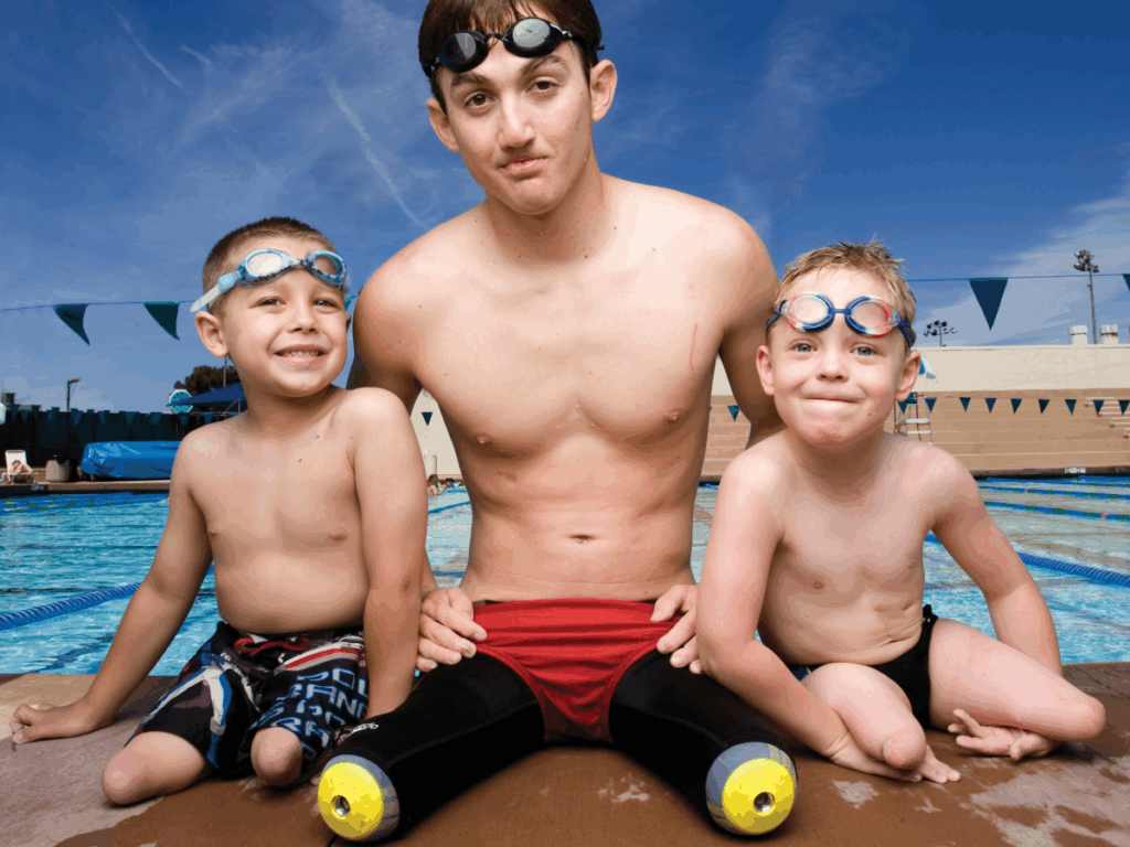 Young male swimmer with bilateral leg prosthetics sits poolside with two young boys, all wearing swim goggles and ready to swim