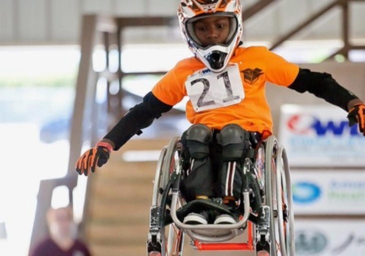 Young athlete wearing an orange jersey and helmet performs a jump in a wheelchair during a WCMX competition