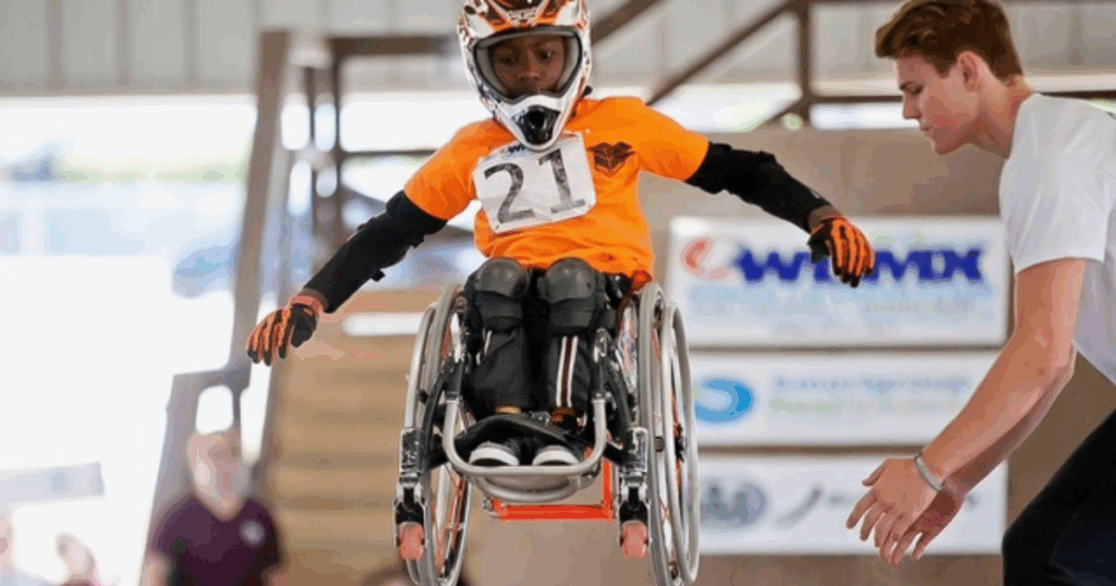 Young athlete wearing an orange jersey and helmet performs a jump in a wheelchair during a WCMX competition
