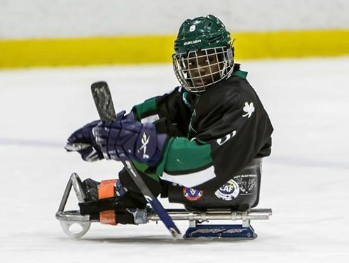 Athlete in full hockey gear competes in a sled hockey game, gripping a stick and moving across the ice