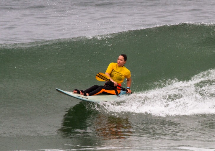 Alana Nichols surfing a wave on an adaptive board, wearing a yellow competition jersey and holding paddle blades