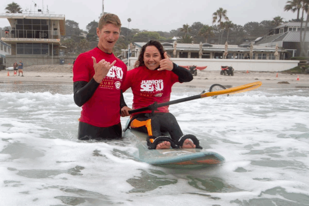 Alana Nichols smiling with a fellow surfer in the ocean, both wearing red Junior Seau Adaptive Surf Program shirts.