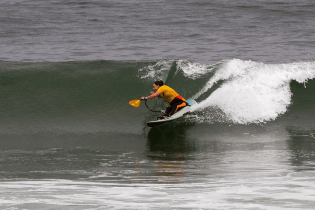 Alana Nichols surfing a wave on an adaptive board, wearing a yellow competition jersey and holding paddle blades