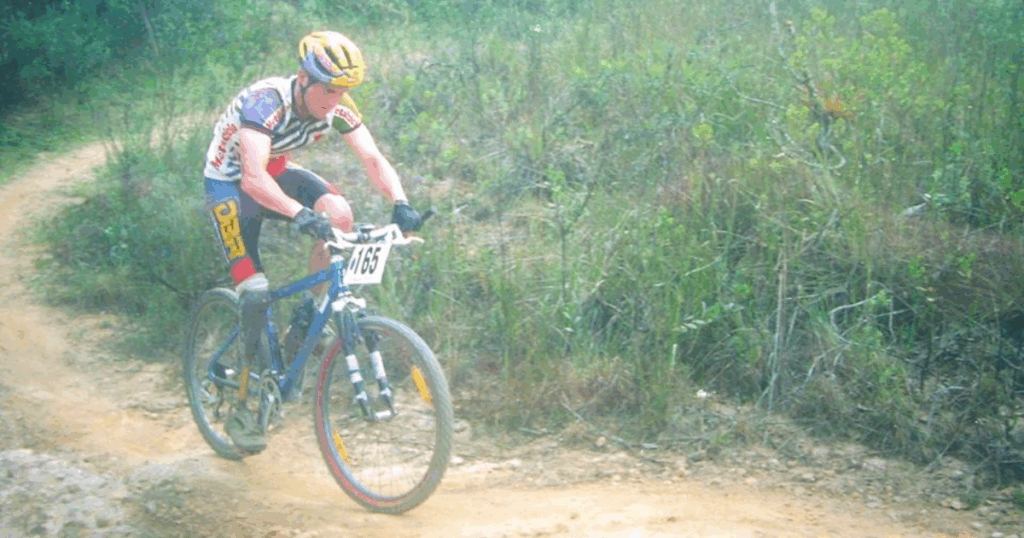 Andre Szucs rides a mountain bike on a dirt trail during an early competition, wearing a yellow helmet and race bib #165