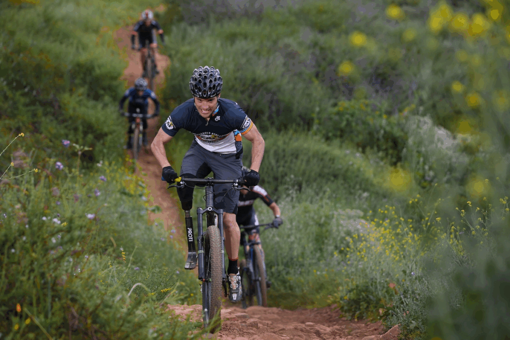 Andre Szucs powers uphill on a rugged dirt trail using his prosthetic leg, leading a group of mountain bikers through the course