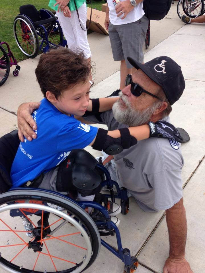 A young boy in a wheelchair hugs an older man, Mike Box, highlighting connection and mentorship in adaptive sports.