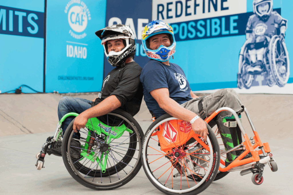 Two athletes in custom wheelchairs sit back-to-back at a skatepark, smiling after a WCMX clinic.