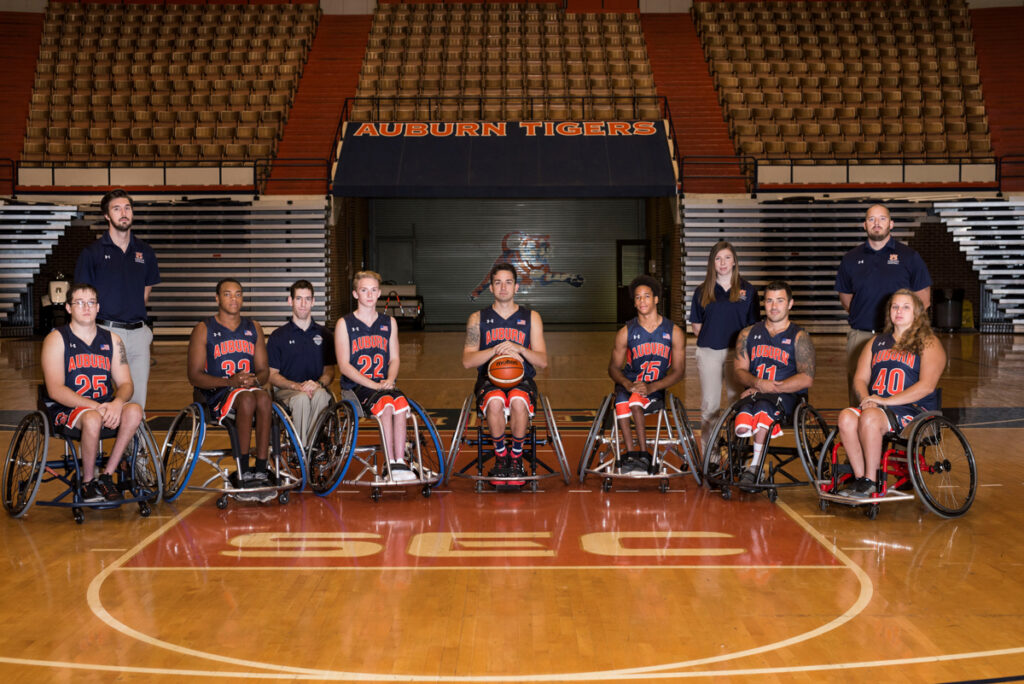 Wheelchair basketball team posed on a court with players in navy jerseys and staff standing behind them.
