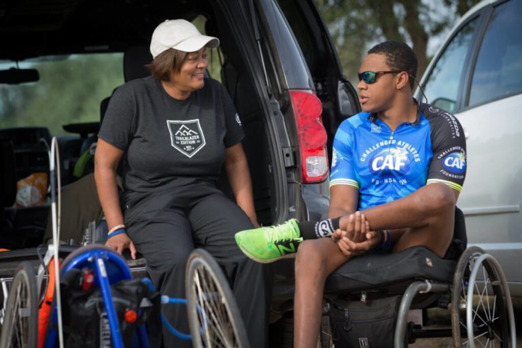 Athlete in a wheelchair wearing a blue CAF jersey talks with a woman beside an open vehicle.