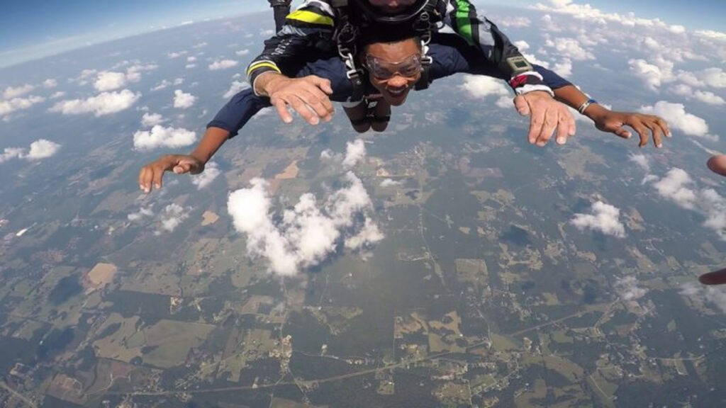 Tandem skydivers freefalling with arms extended above fields and clouds.