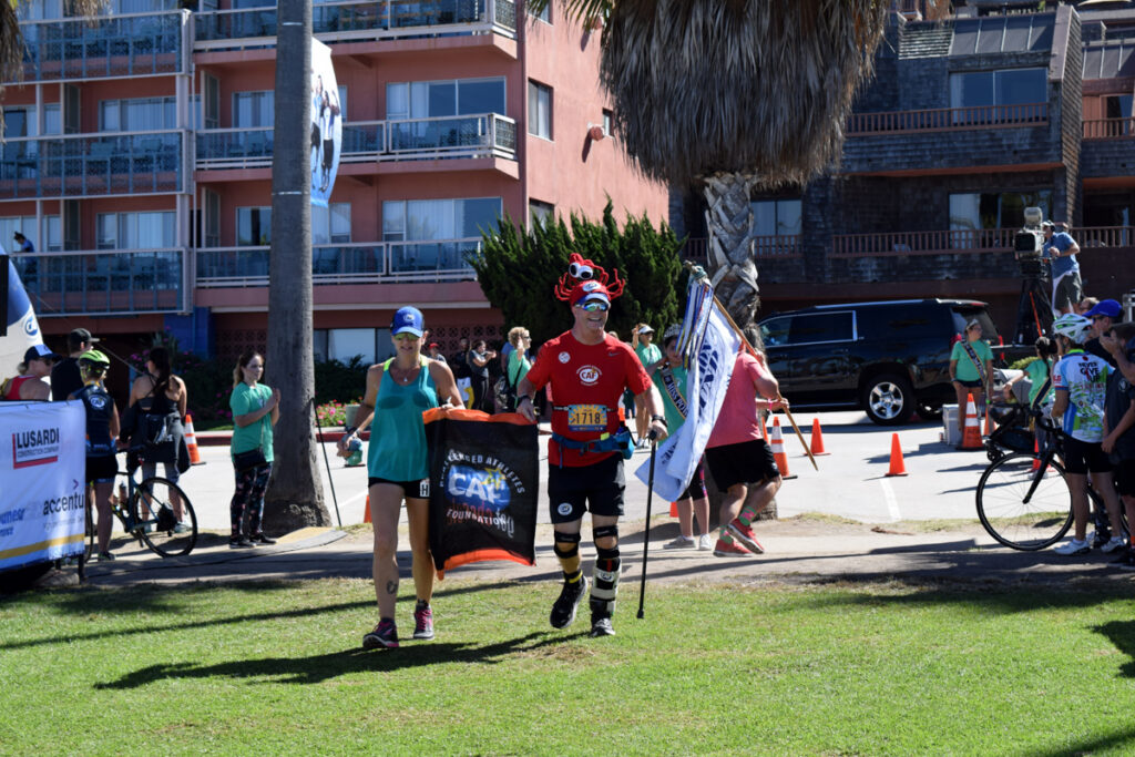Two people in athletic gear run through a grassy area, surrounded by onlookers and buildings in the background. One holds a flag.