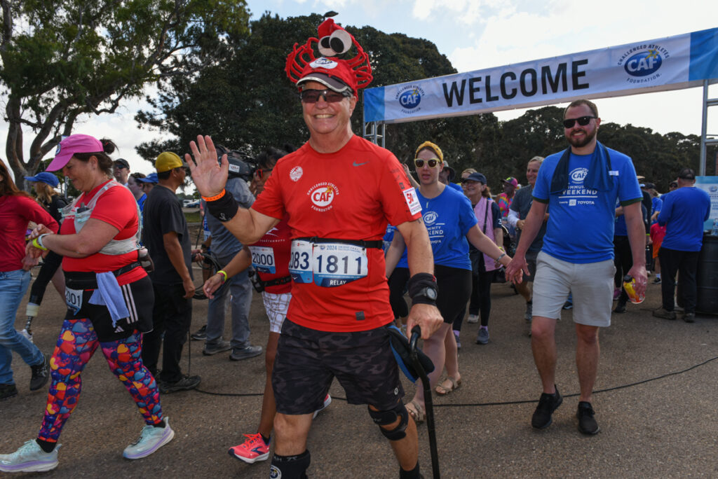Participants in a race walk under a welcome banner, with one wearing a red crab hat and using a cane.